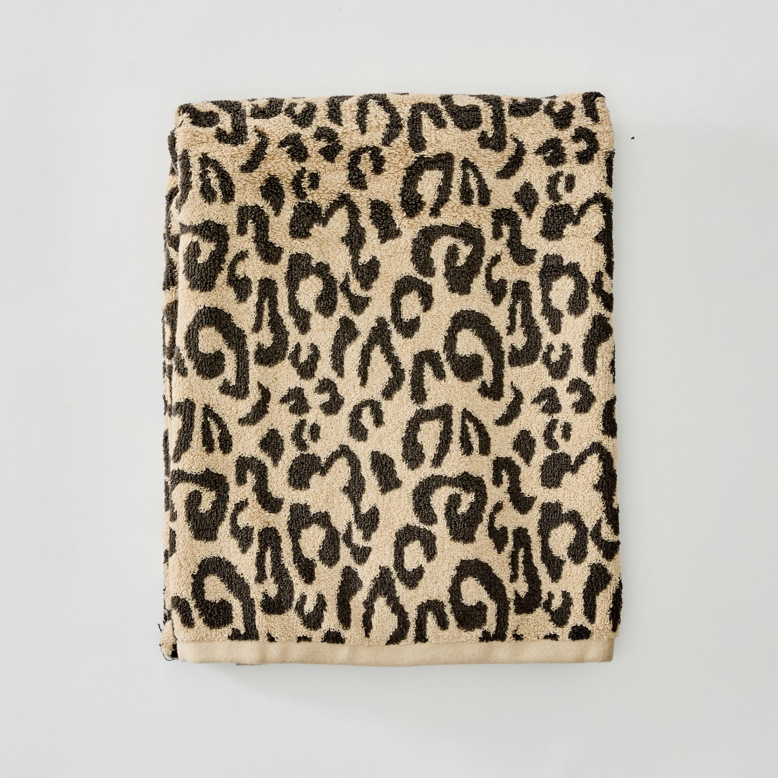 A folded cotton towel with a classic leopard print pattern in black on a warm beige background, featuring a solid beige border along the bottom edge, photographed flat against a light gray background.