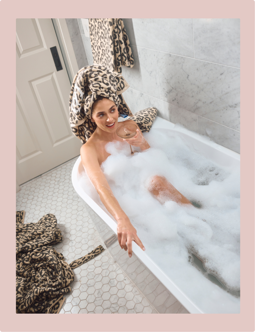 A woman relaxes in a bubble-filled bathtub with a leopard-print towel wrapped around her head and matching towels on the floor, set in a modern tiled bathroom.