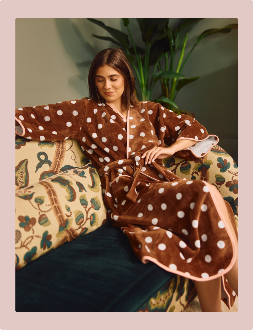 A woman reclining on a patterned sofa wearing a plush brown robe with white polka dots and pink trim, set against a dark green background with a plant.