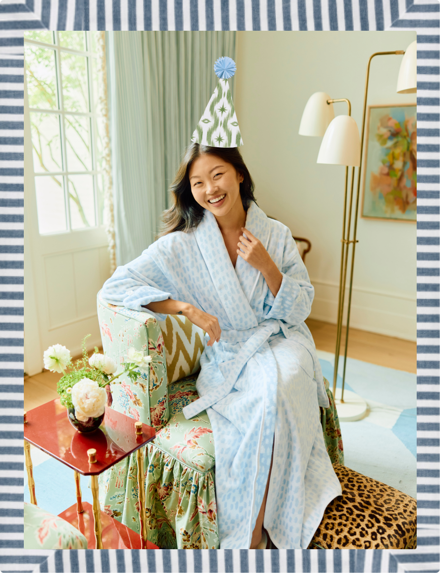 A woman wearing a light blue patterned bathrobe and a green-and-white party hat sits smiling on a floral armchair in a bright room. The photo is framed by a blue-and-white striped border.