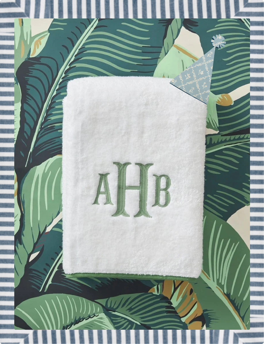 A white plush towel featuring green embroidered initials 'AHB', styled on a tropical green leaf patterned background with a striped border.