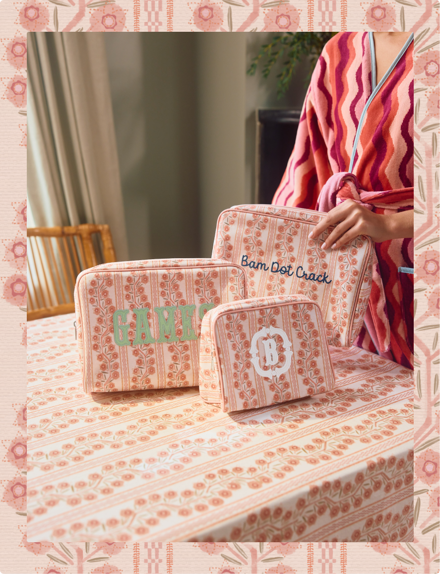 Three matching fabric cosmetic or storage pouches with a pink and peach floral stripe pattern are arranged on a table. One pouch has the word "games" in green, one has "Bam Dot Crack" in navy, and one has a circular monogram logo with "B".