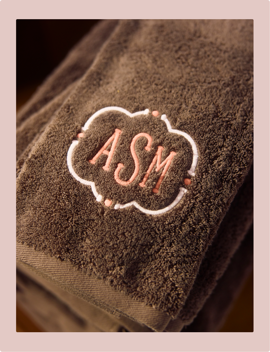 A close-up of a folded brown towel with a monogrammed patch reading 'ASM' in pink and white embroidery, set against a soft pink background.