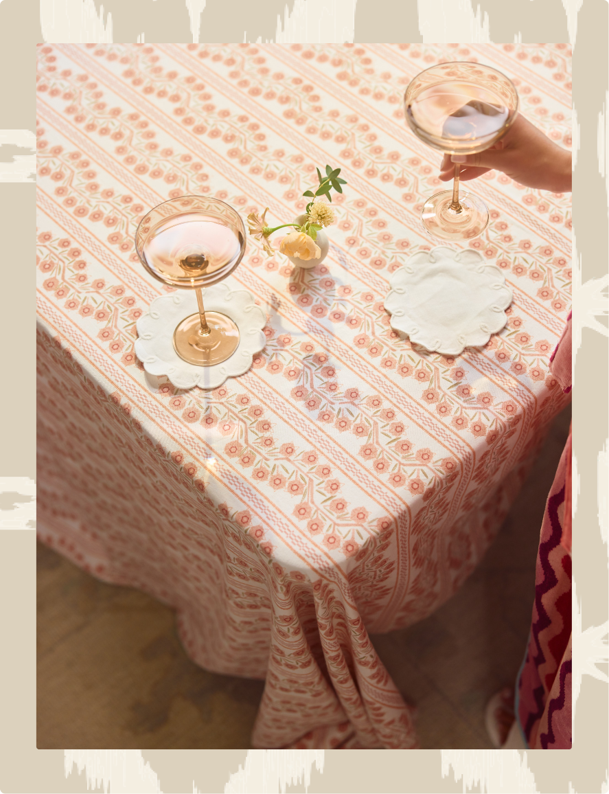 A peach and white patterned tablecloth draped over a table, styled with two champagne glasses, white coasters, and a small floral centerpiece. A hand is holding one of the champagne glasses, and a portion of a person's patterned dress is visible on the right side.