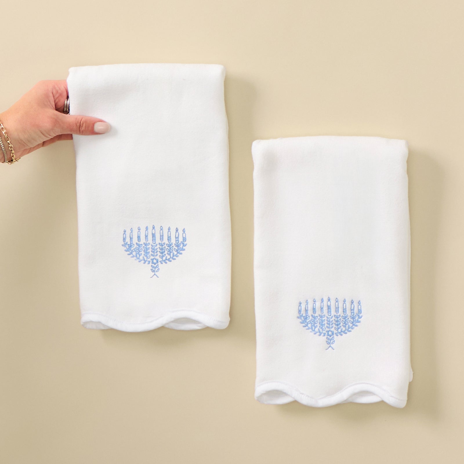 Menorah scalloped powder bath towels, a pair with blue menorah embroidery, held on a cream backdrop