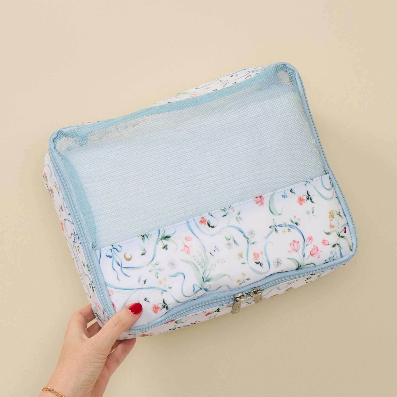 A hand with red painted nails holds a rectangular travel organizer bag with a floral print and light blue mesh pocket, set against a solid beige background.