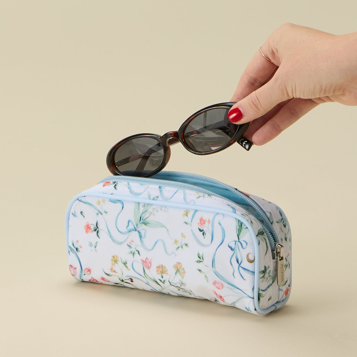 A hand with red nail polish holds a pair of dark sunglasses above a zippered cosmetic pouch. The pouch has a white background with a delicate pastel floral print and light blue wavy line details, set against a solid beige background.