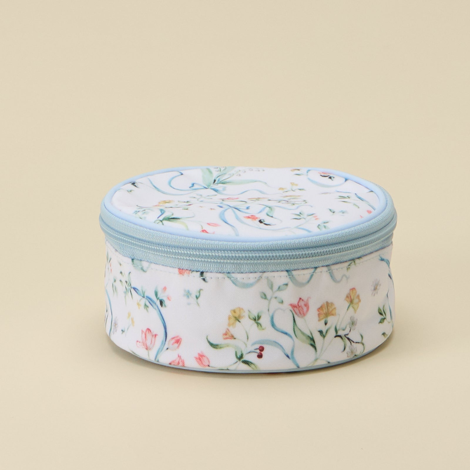 A round zippered cosmetic bag with a white background and a delicate multicolor floral pattern, featuring a light blue zipper and trim, set against a solid beige background.