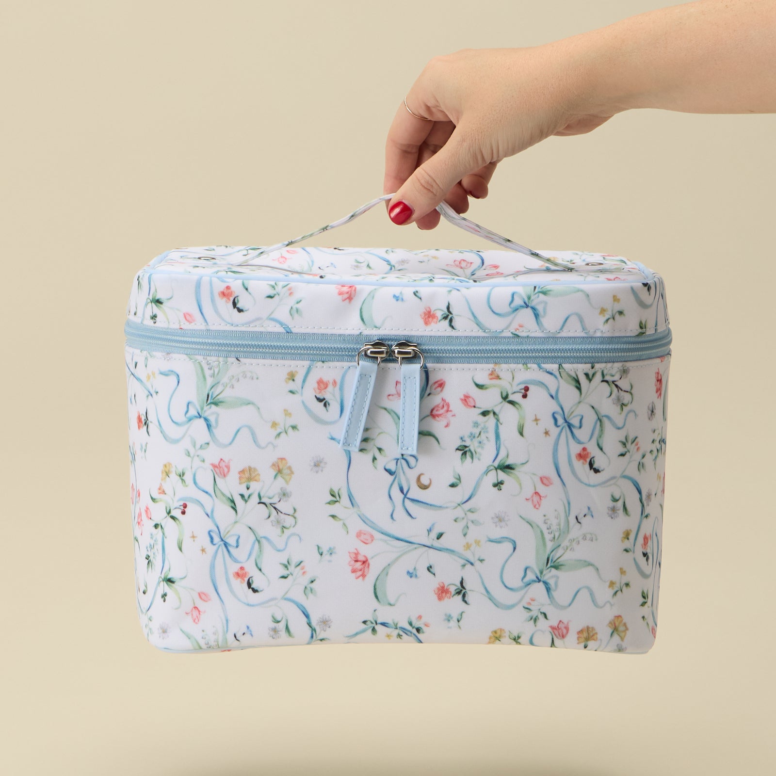 A hand with red nail polish holds the top handle of a square zippered cosmetic bag. The bag has a white background with a delicate blue, pink, and green floral and vine pattern, and a light blue zipper.