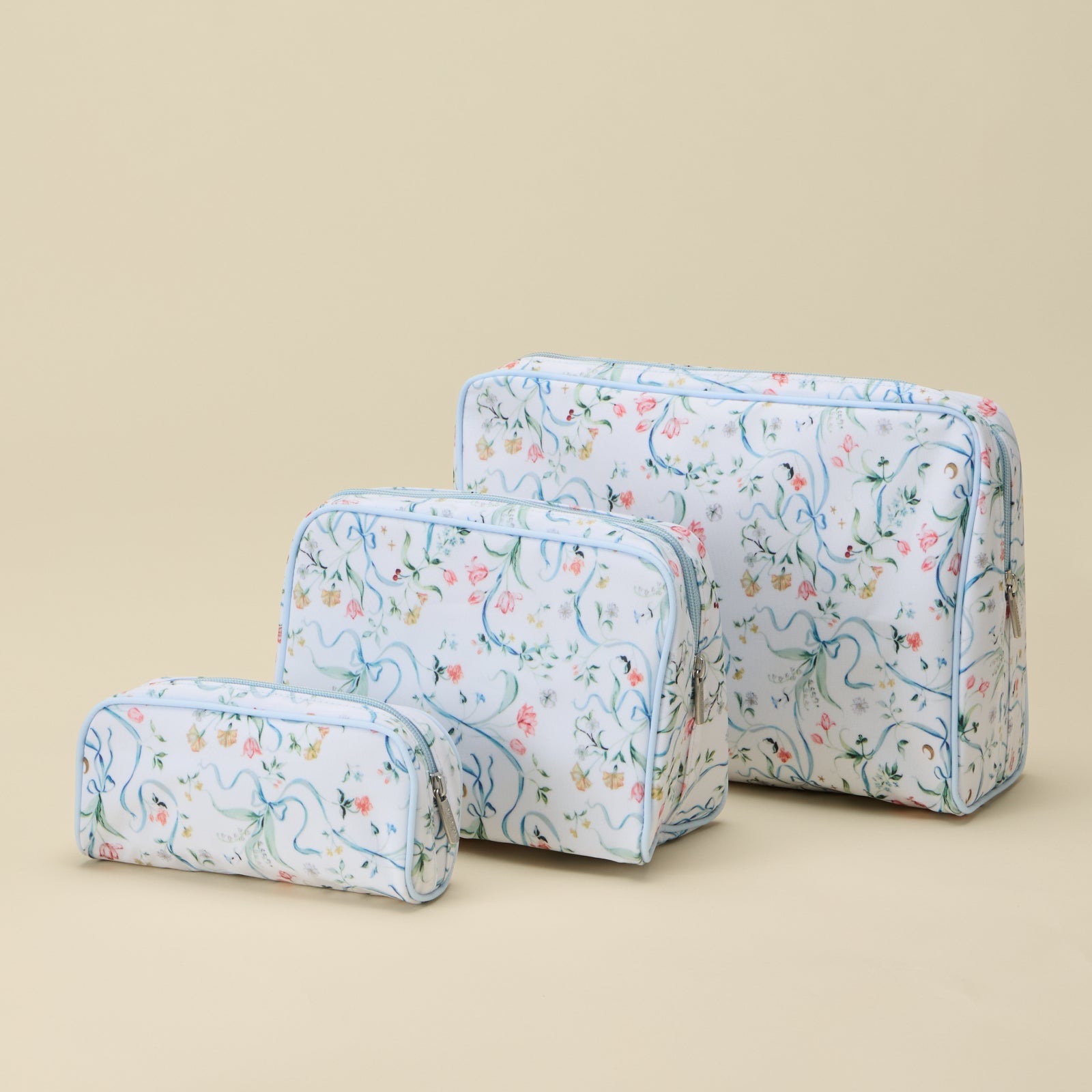 Three rectangular zippered cosmetic or travel pouches in a small-to-large size set, featuring a delicate multicolor floral and vine pattern on a white background with light blue edging, arranged on a solid beige background.