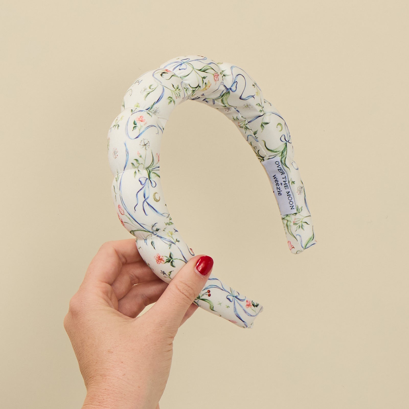 A hand with red nail polish holds a padded fabric headband featuring a white background with a delicate blue, green, and pink floral vine pattern. A white fabric tag is visible on the inside of the headband, reading 'DAMASK HEADBANDS'.