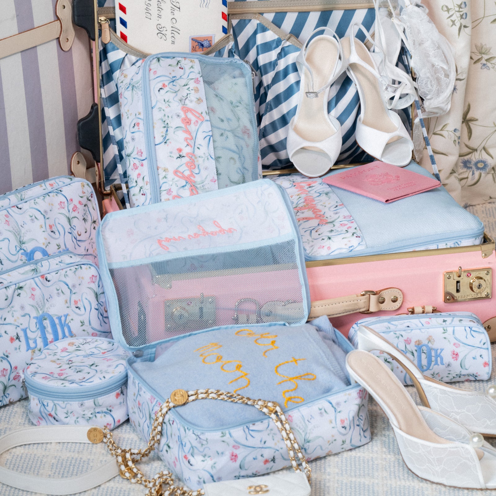 A coordinated set of pastel travel packing cubes and small zip pouches with a light blue floral print, arranged around a pink suitcase. White heeled sandals, a striped bag, and other travel accessories are visible in the styled scene.