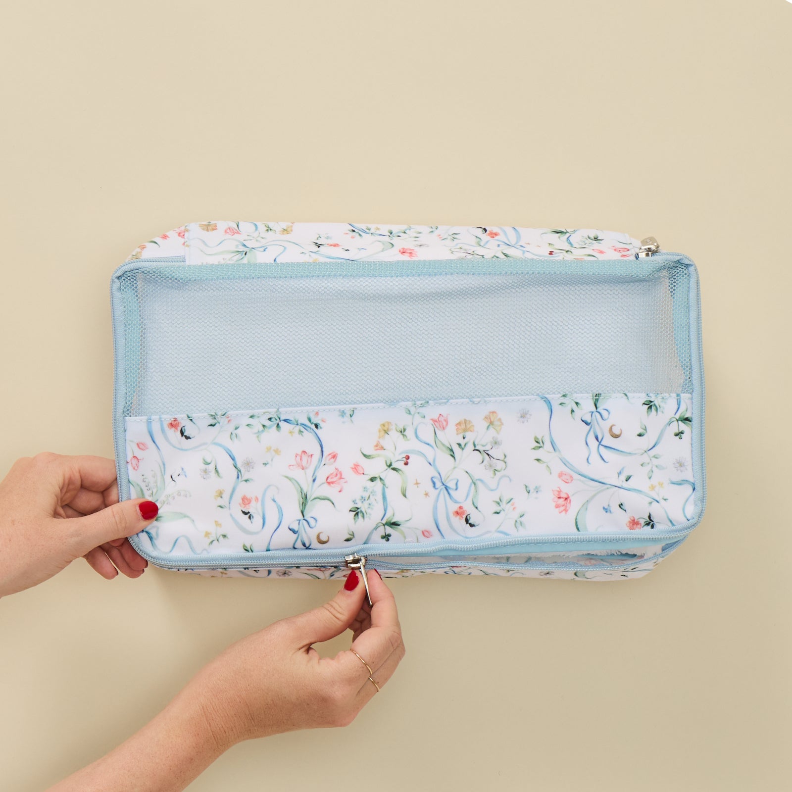 A light blue and white floral toiletry bag with a mesh window, held open by two hands with red nail polish against a solid beige background.