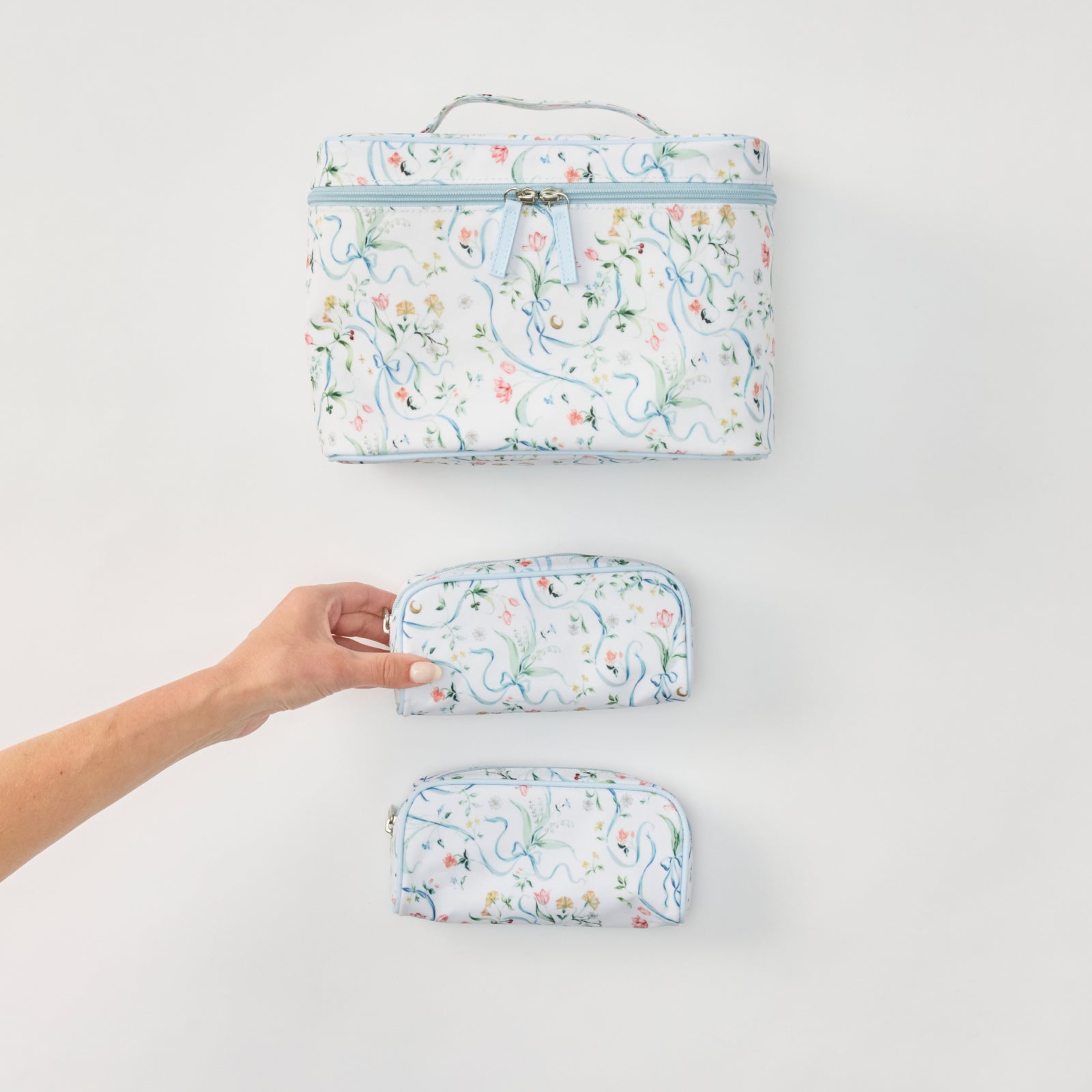 Three matching fabric zippered pouches or small cases with a white background and a delicate multicolor floral print, arranged vertically on a light gray surface. A human hand is holding the middle case.