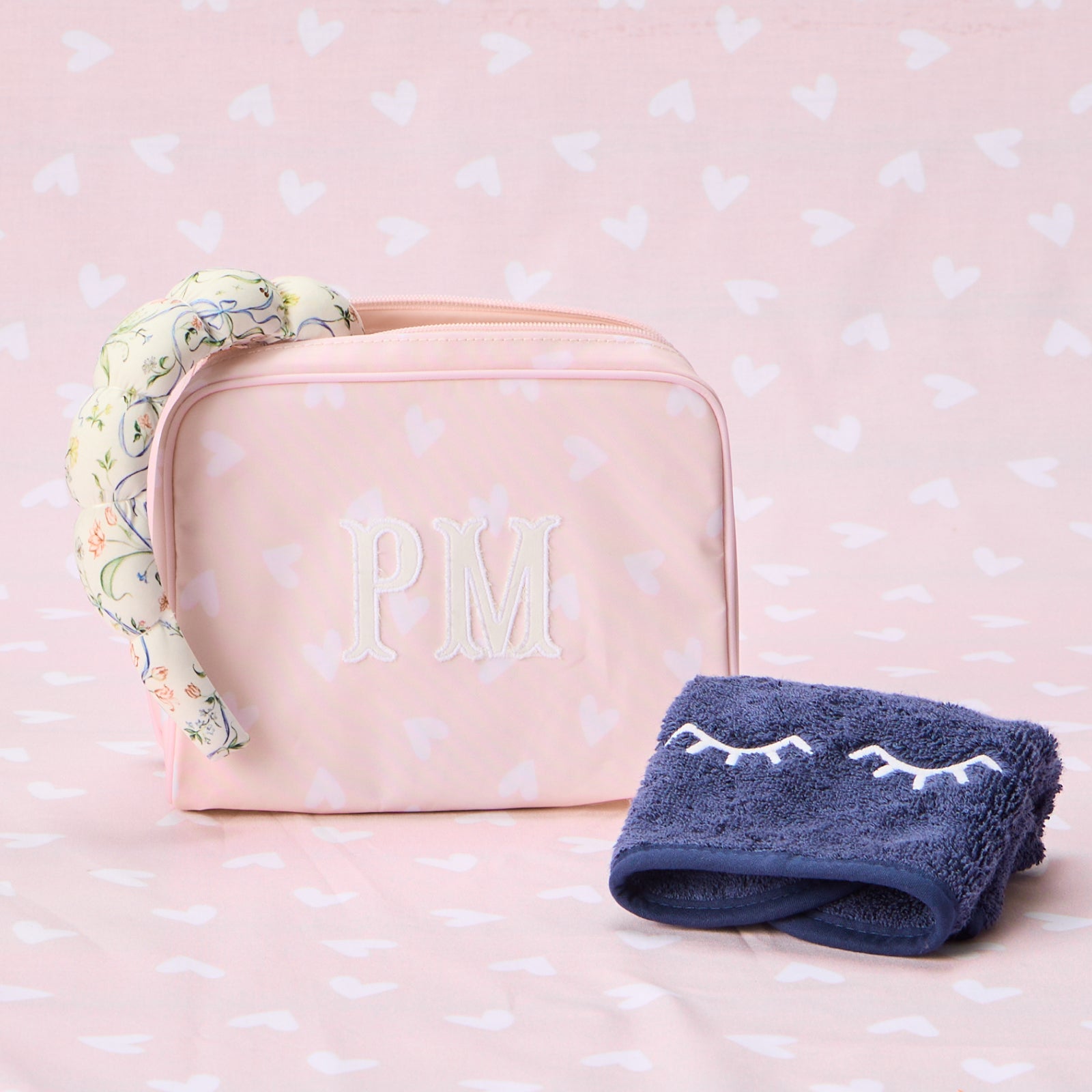 A pink heart-patterned medium patterned toiletry bag with "PM" monogrammed in white, next to a matching navy eye mask.