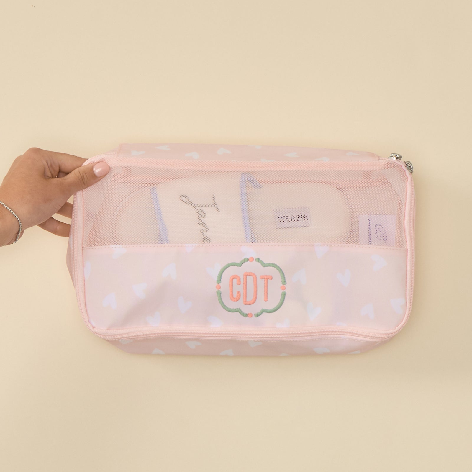 Top-down view of a rectangular, zippered Medium Packing Cube in a "Tossed Pink Hearts" print. The cube features a clear mesh top section and a pale pink lower section patterned with white hearts. The lower front panel is embroidered with the initials "CDT" in green, cursive thread inside a decorative scalloped border. A pair of beige slippers is visible inside through the mesh, and a hand is shown holding the edge of the cube against a solid, light-toned background.