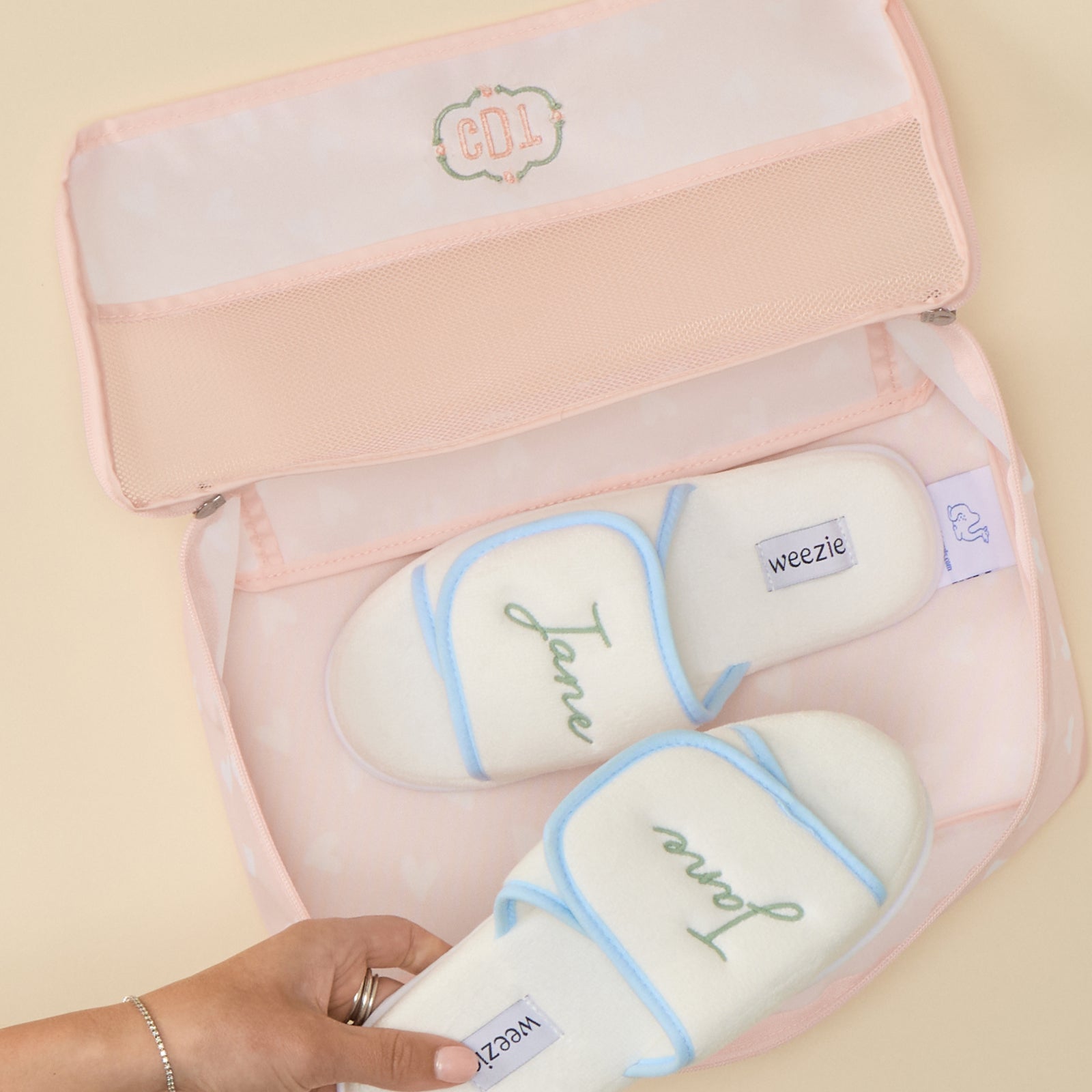 Top-down view of an open Medium Packing Cube in the "Tossed Pink Hearts" print. The cube features a pale pink lower section patterned with white hearts, and the open clear mesh lid has the initials "CDL" embroidered in green thread inside a decorative scalloped border. Inside the cube, two cream-colored slippers with blue trim are packed, each embroidered with the name "Jane" in green cursive script. A hand is holding the edge of the cube against a solid, light-toned background.