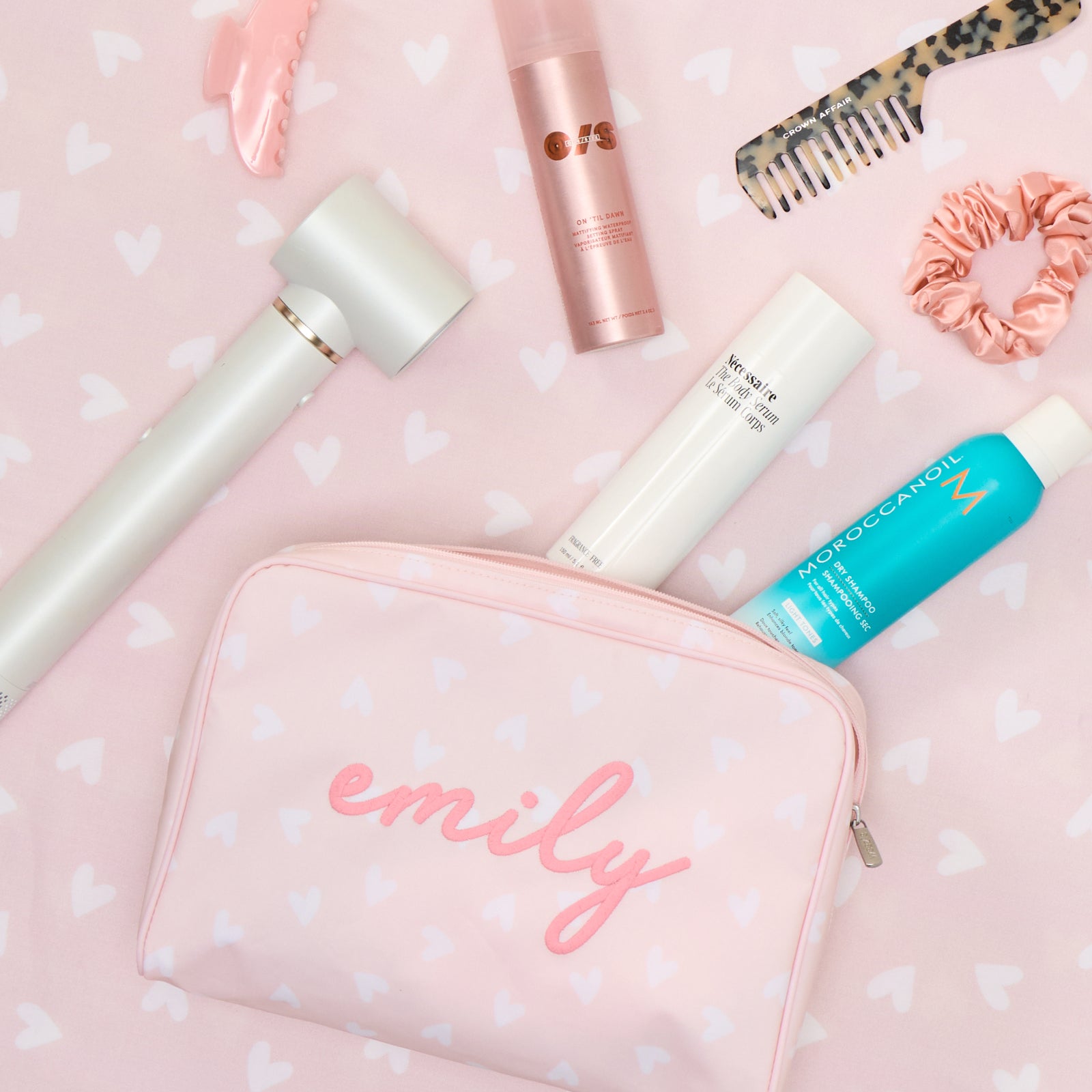 A personalized pink heart-patterned toiletry bag labeled "emily" surrounded by various beauty products and accessories.