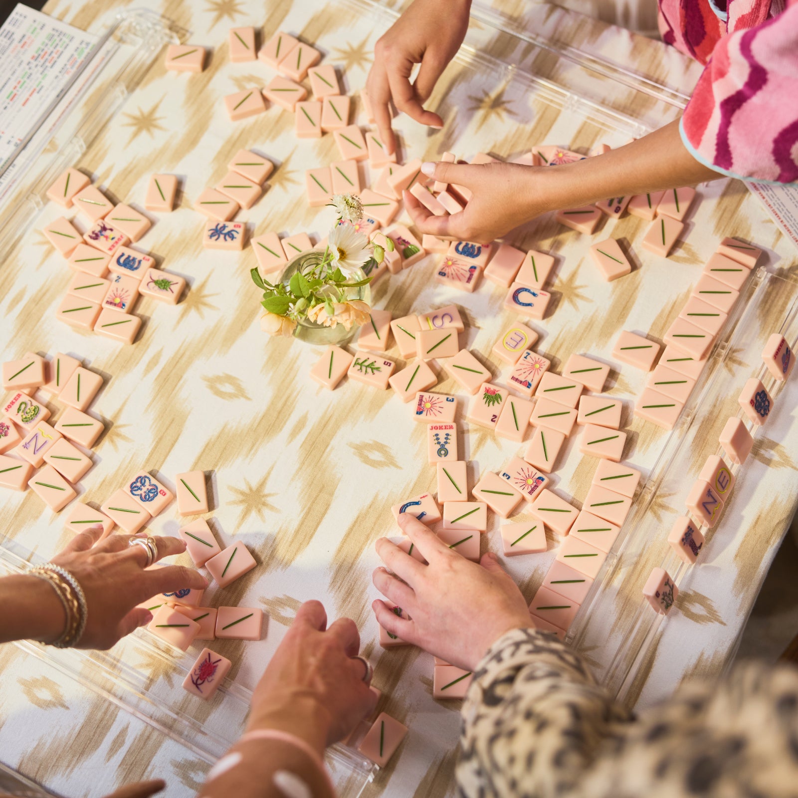Several people's hands are visible arranging pastel pink and multicolored Mahjong tiles on a patterned table runner in a bright indoor setting.