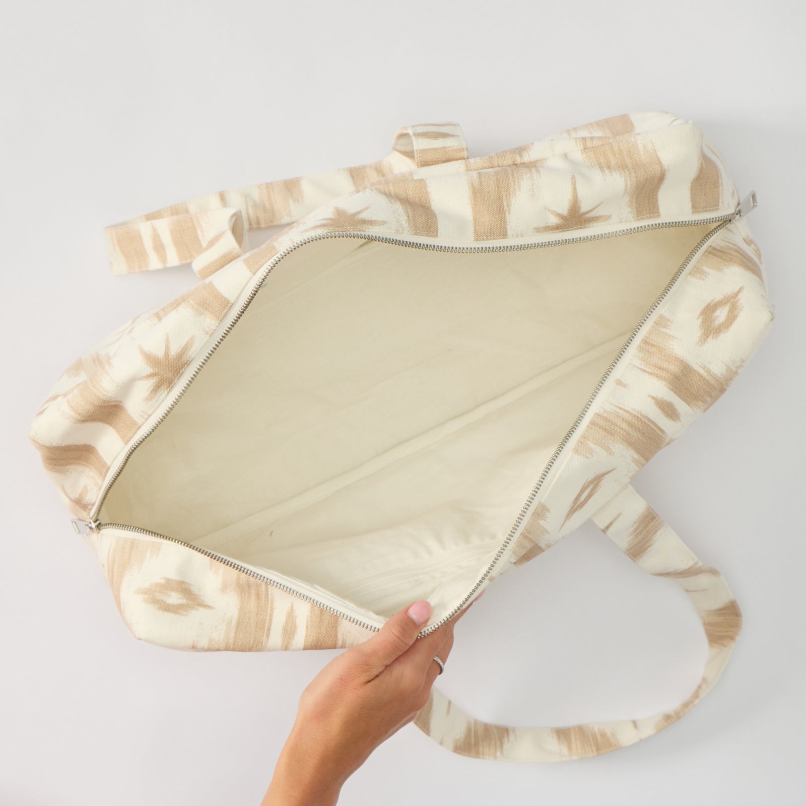 A close-up of a person's hand pulling open a wide fabric Mahjong bag to show its empty, light-colored interior. The bag and its straps are decorated with a tan and cream ikat design.