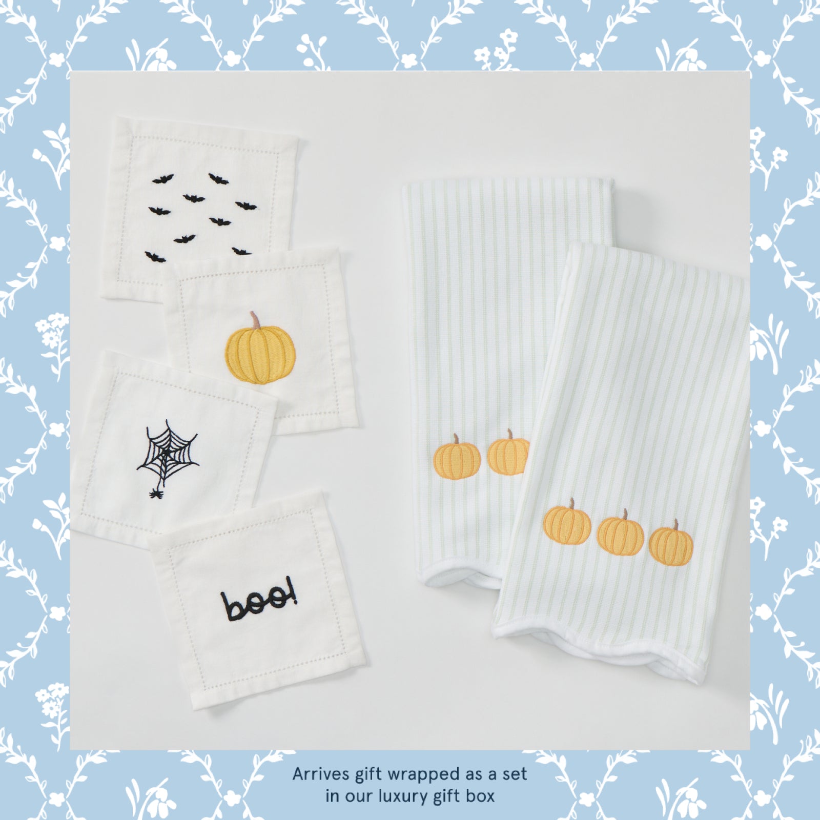 A set of white Halloween-themed baby textiles, including two ribbed square cloths with embroidered pumpkins and three smaller square cloths with embroidered bats, a spider web, and the word 'boof!' in black, displayed on a white background with a blue floral border.