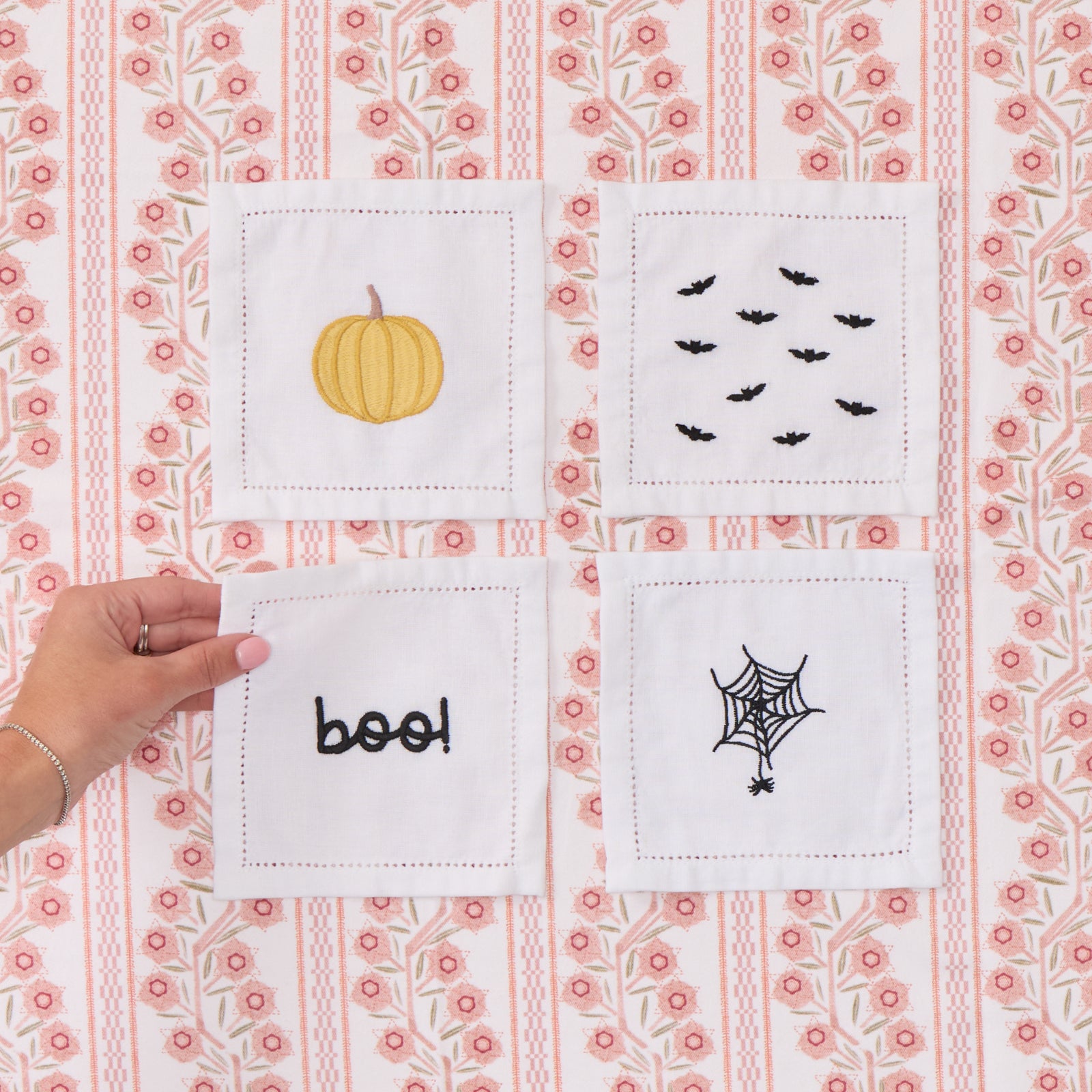 Four white cocktail napkins with Halloween embroidery: bats, pumpkin, boo, and spiderweb.