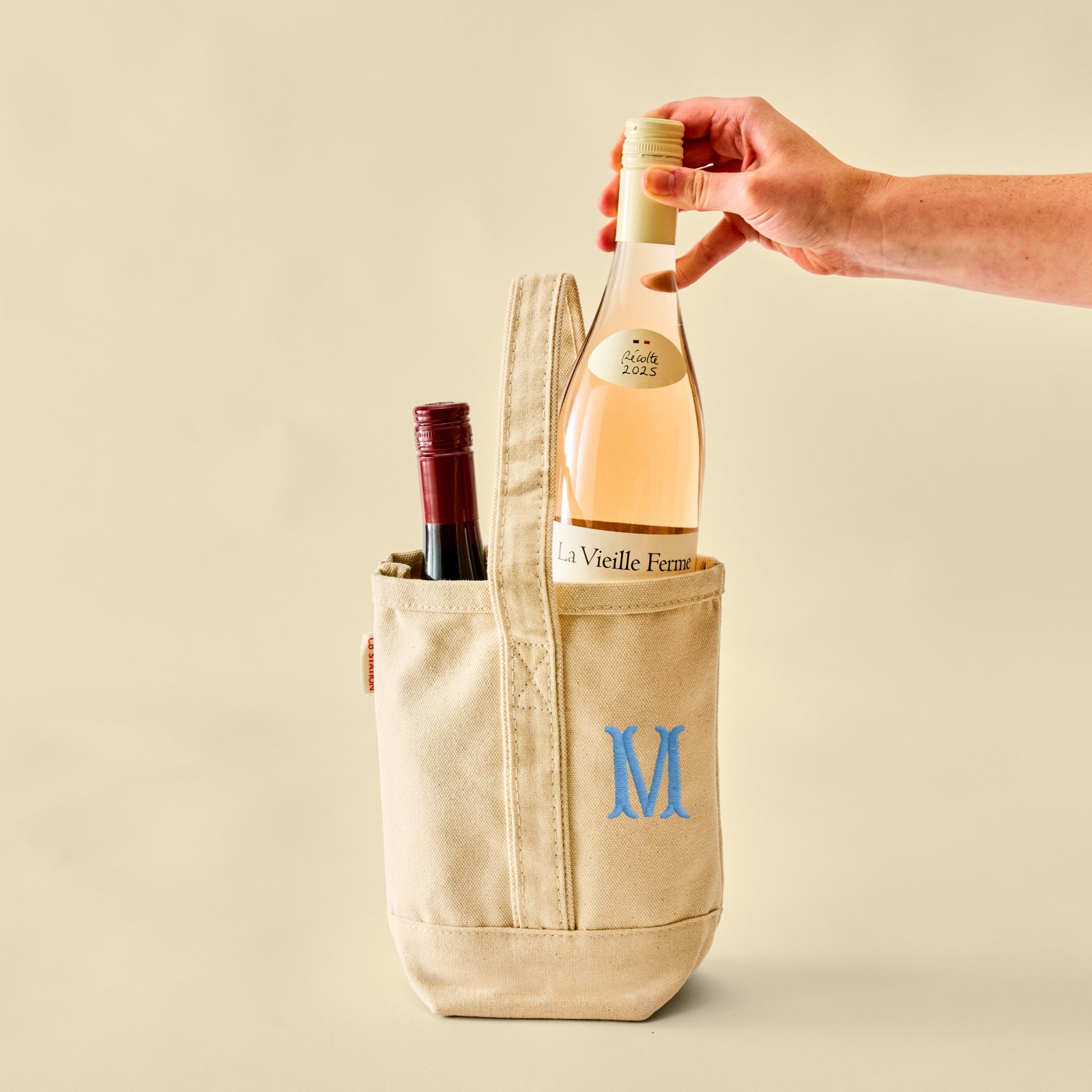 A beige canvas wine tote bag with a blue embroidered letter 'M' on the front pocket, holding two wine bottles. A hand is reaching in to grab the rose wine bottle from the front pocket.