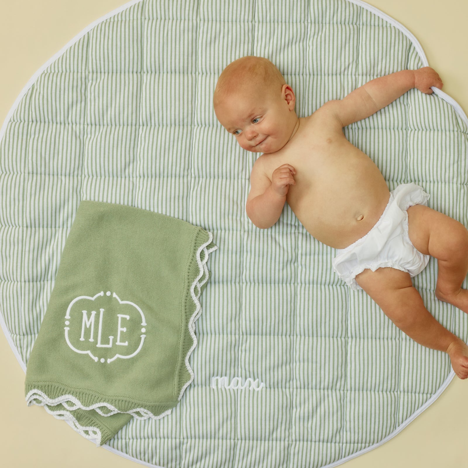 Baby lying on a round green striped mat next to a green scalloped blanket with MLE monogram.