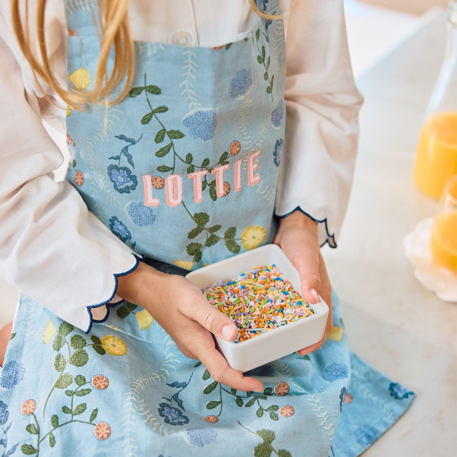 A child in a light blue floral apron embroidered Lottie, holding a bowl of sprinkles.