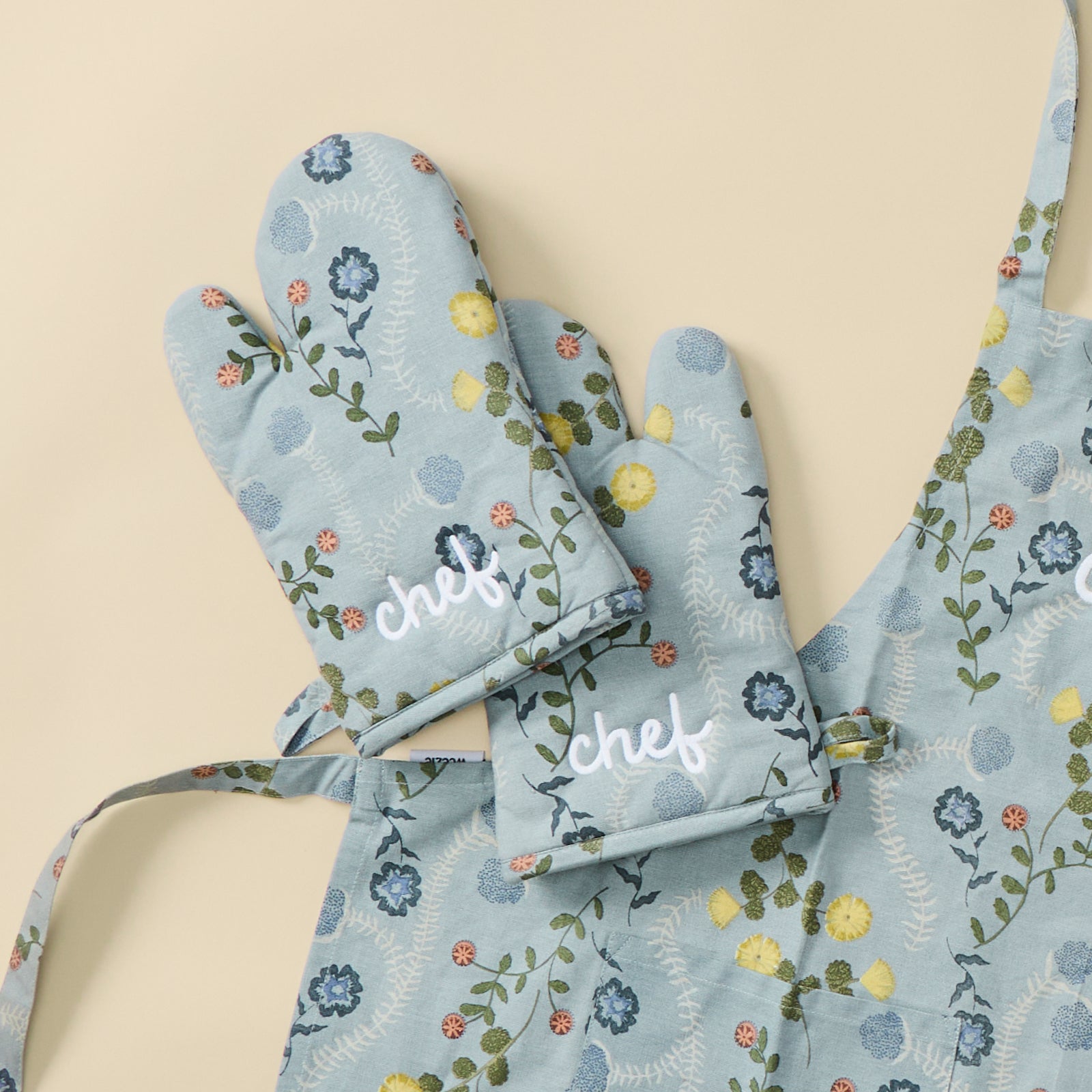A pair of light blue floral oven mitts embroidered chef, laid on a matching apron.