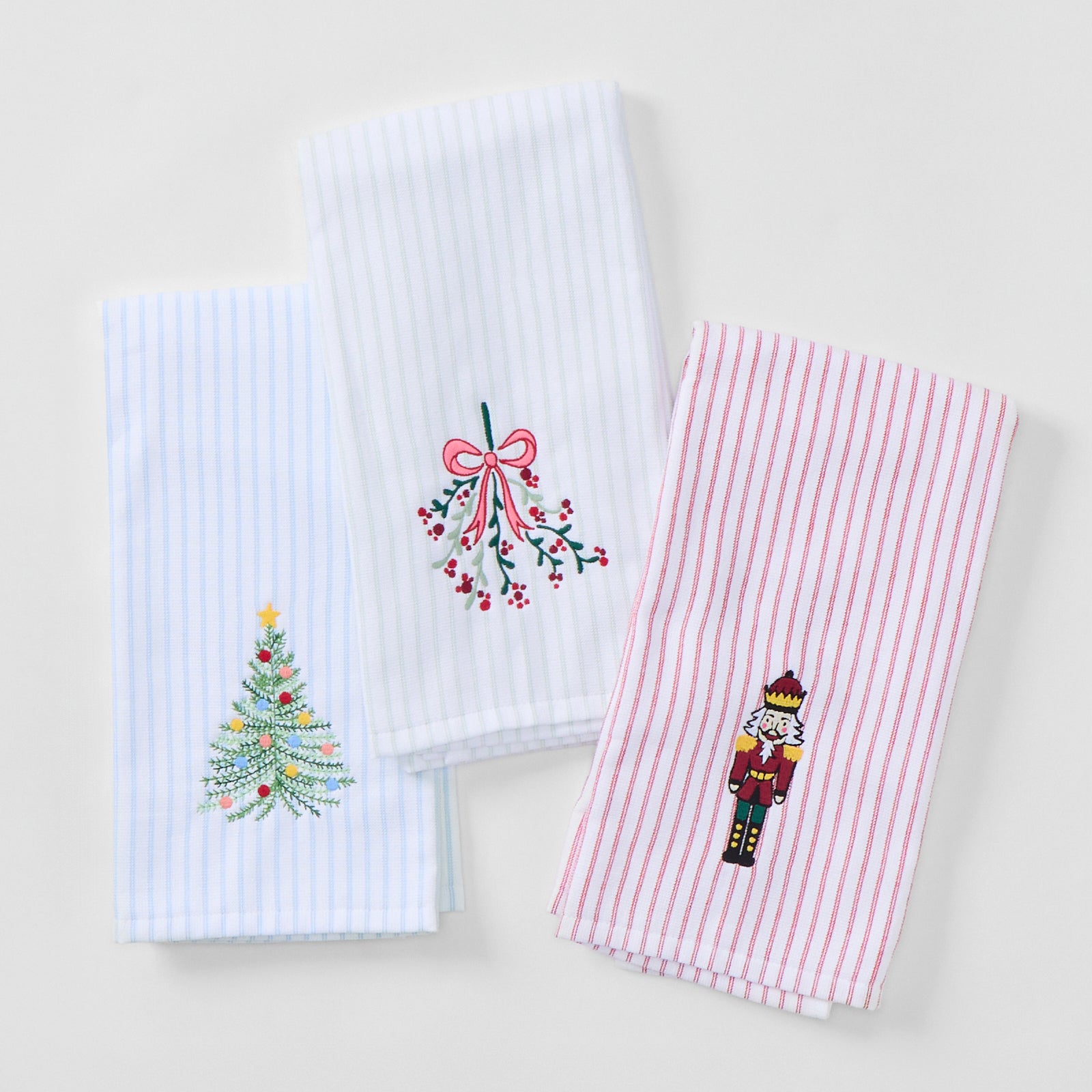Three folded Tidings Trio kitchen towels in pale stripes, embroidered with a Christmas tree, mistletoe bow, and nutcracker