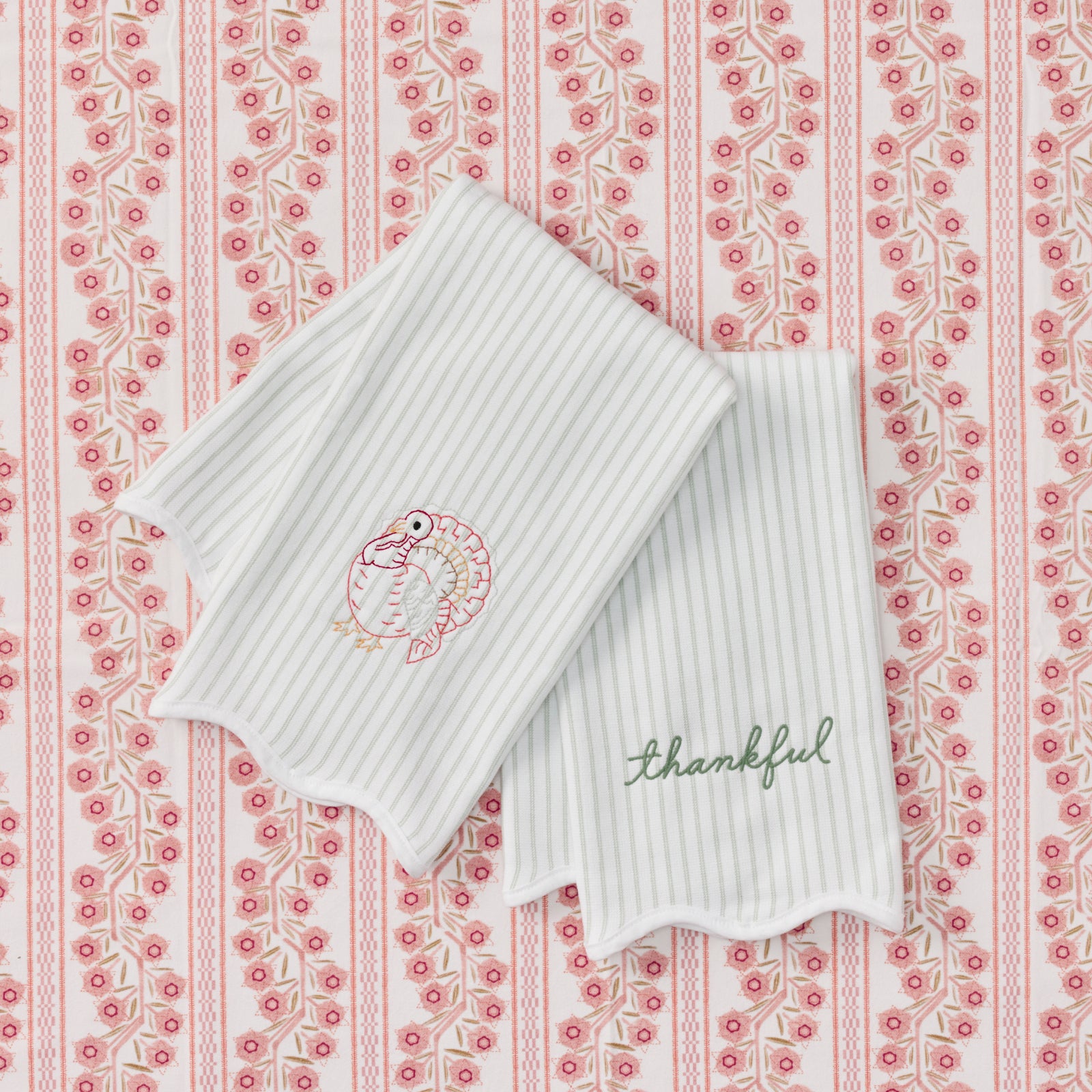 Two green striped kitchen towels with scalloped edges, featuring a pink and green embroidered turkey on one towel and the word 'thankful' in green script on the other, displayed on a pink and white patterned background.