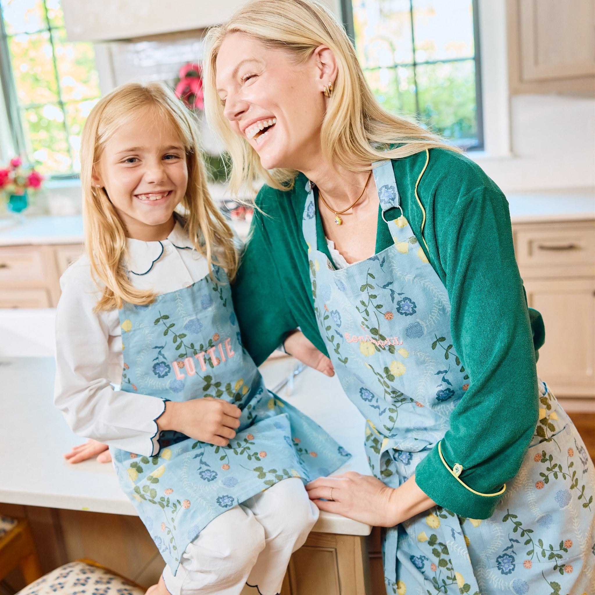 Woman and young girl in matching blue floral print aprons laughing together in a kitchen.