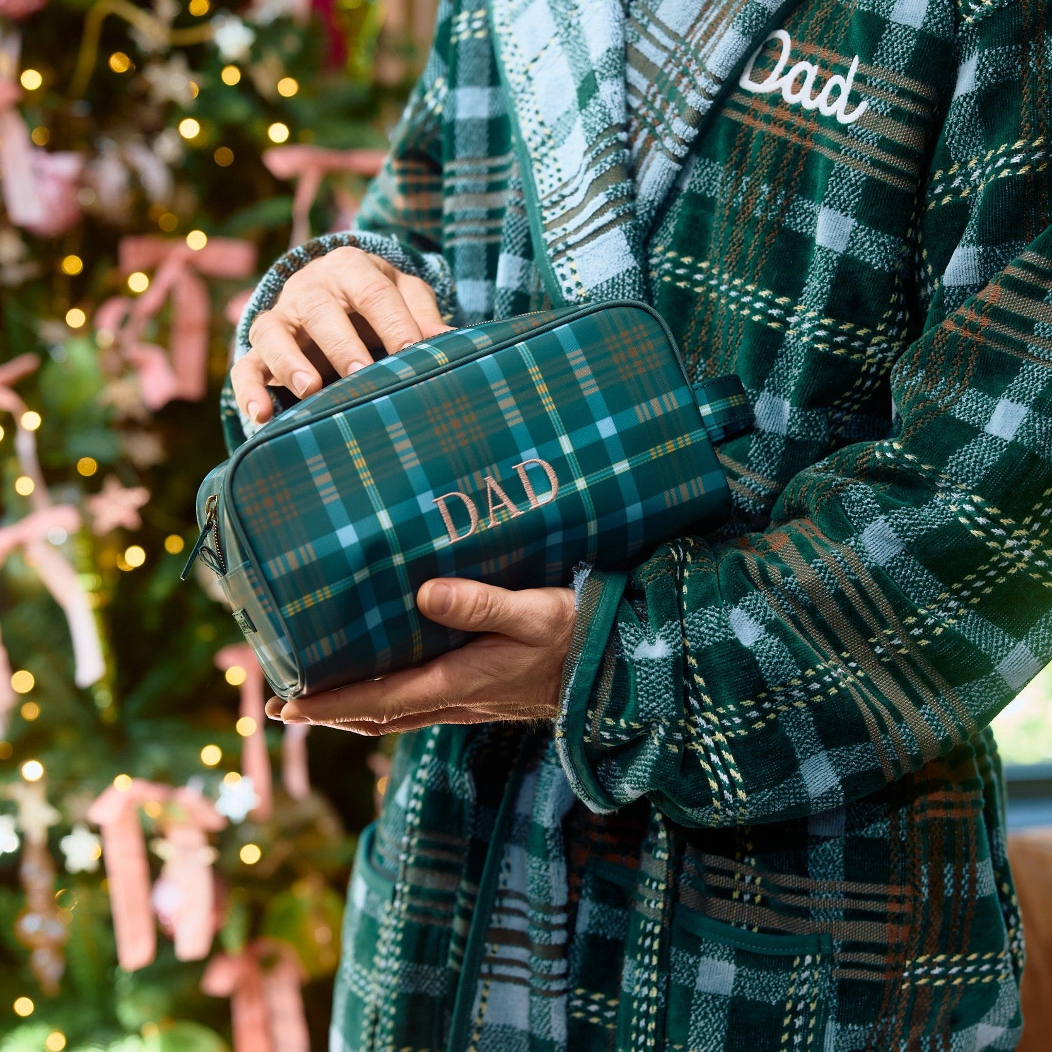 Hands in a green plaid robe holding a matching plaid toiletry bag monogrammed Dad beside a Christmas tree.