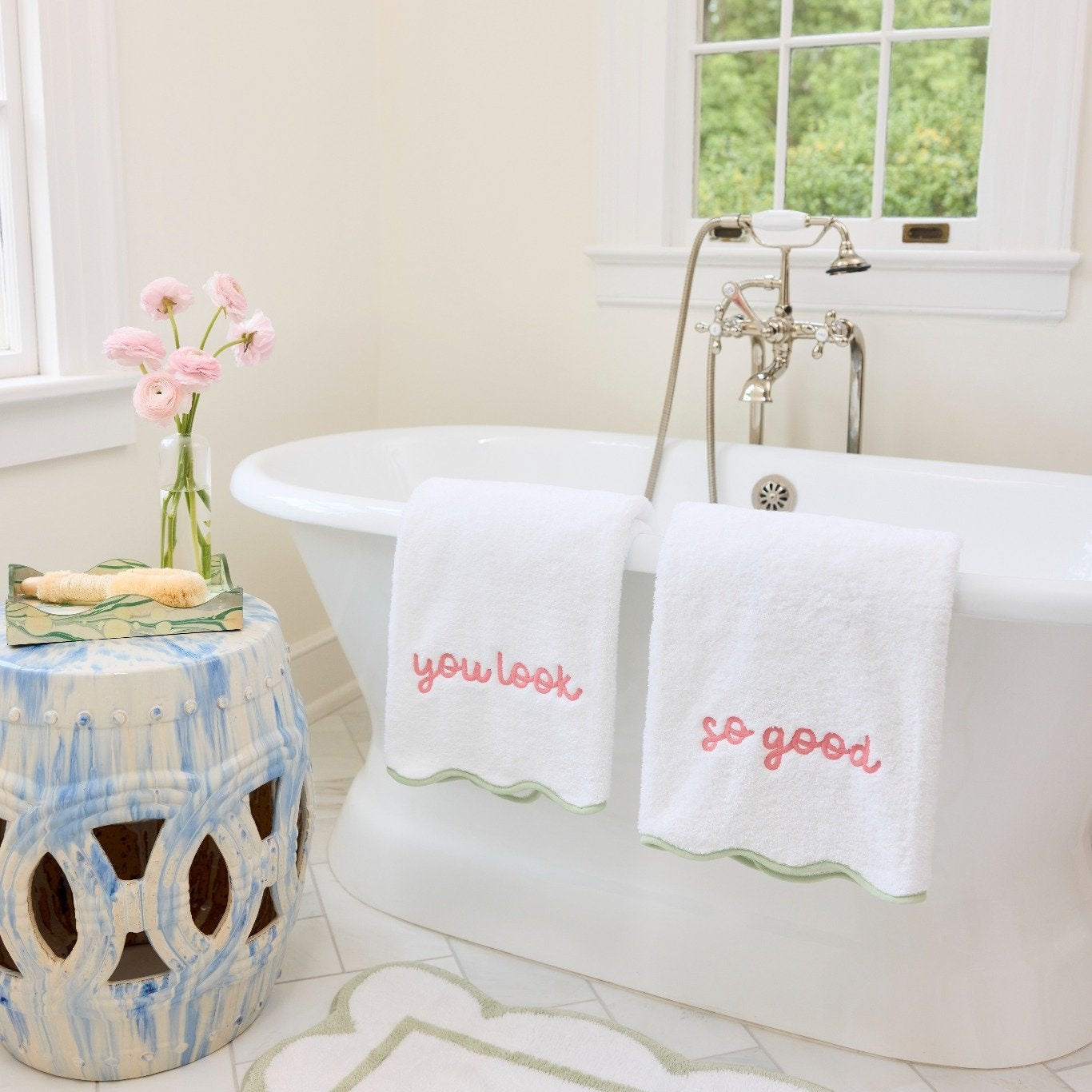 Two white bath towels with pink embroidered text reading 'you look so good' draped over a white freestanding bathtub in a bright bathroom. A patterned blue-and-white side table with pink flowers sits beside the tub, and a white bath mat is on the floor.