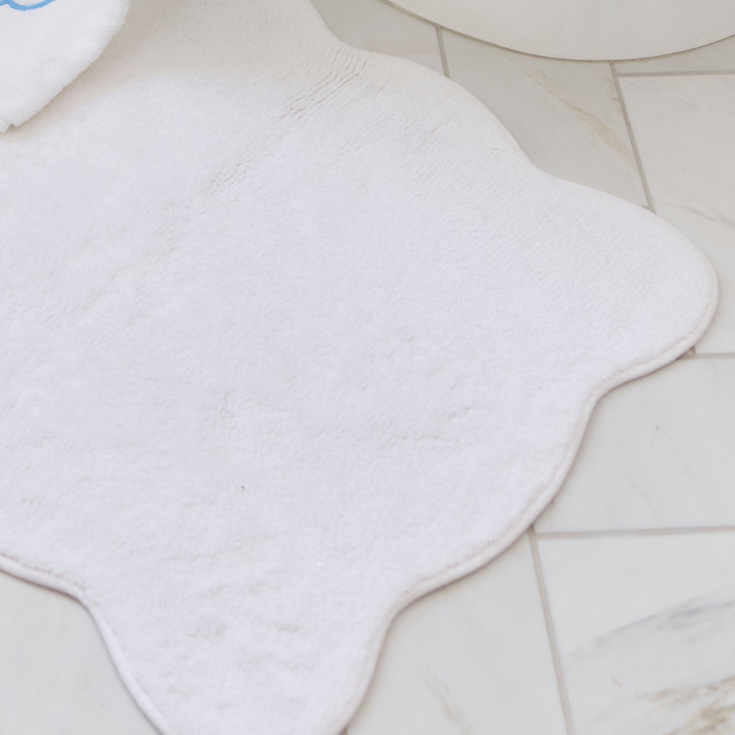 A plush white bath rug with scalloped edges placed on a light-colored marble tile floor.