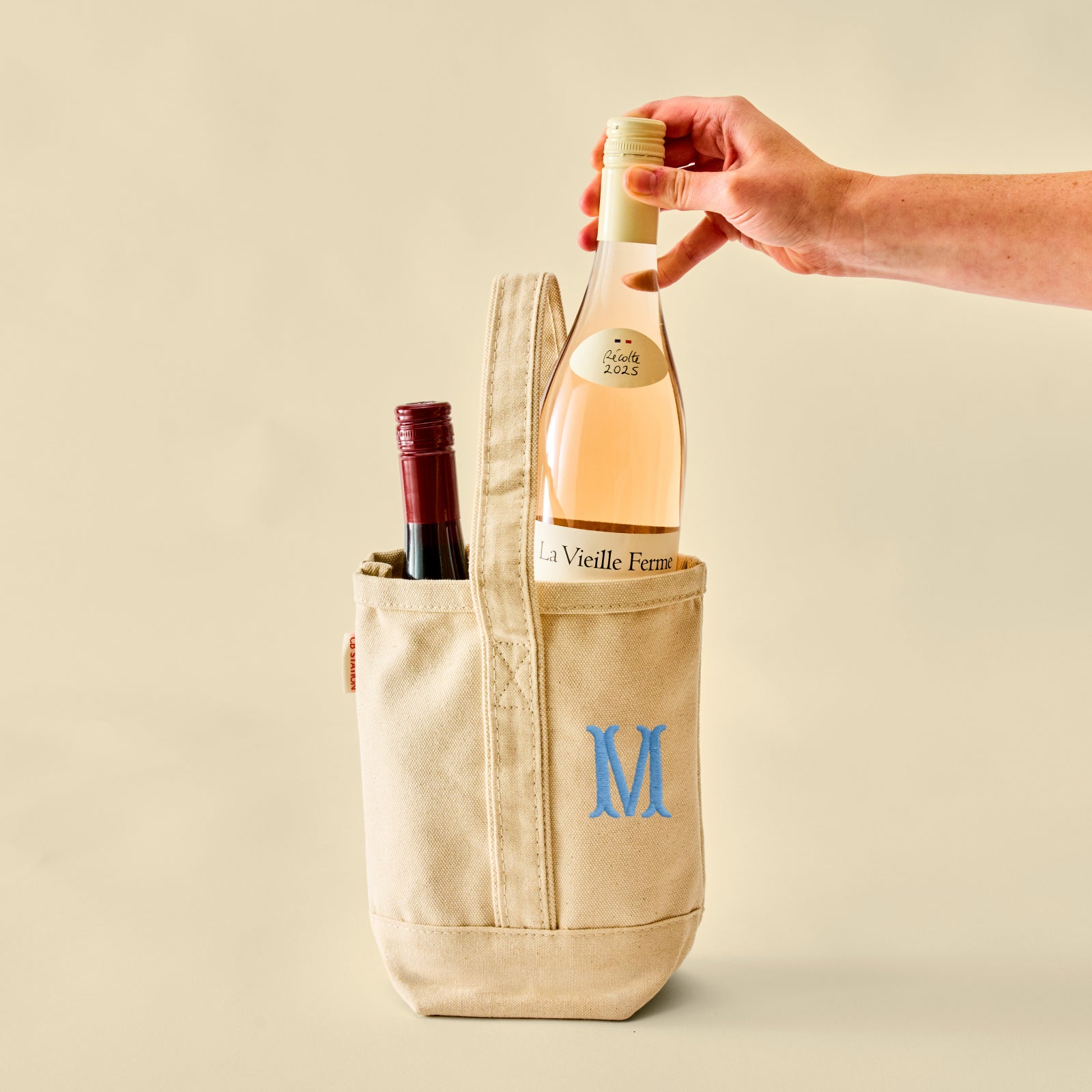 A beige canvas wine tote bag with a blue monogram letter 'M' on the front pocket, holding a bottle of rosé wine and part of another bottle. A hand is reaching in to remove the rosé wine bottle.