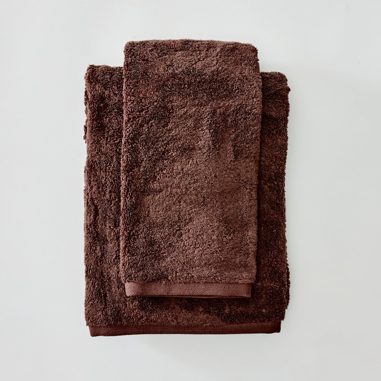 A stack of three dark brown terry cloth towels, including a bath towel and two smaller hand towels, neatly folded on a light gray background.