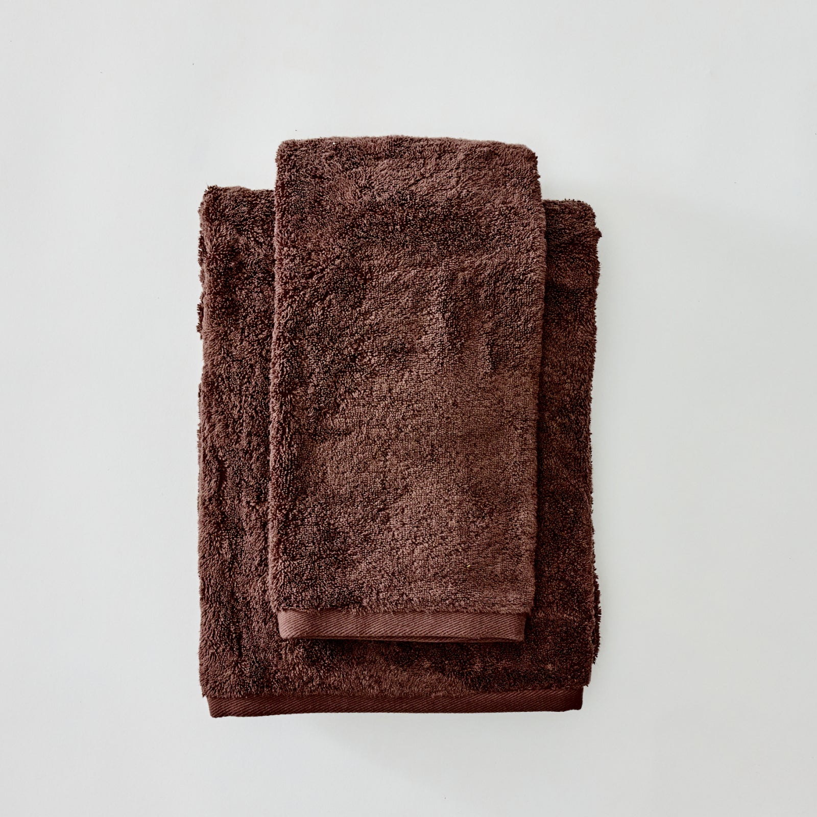 A stack of three folded towels in a rich dark brown color, arranged neatly on a light gray background.