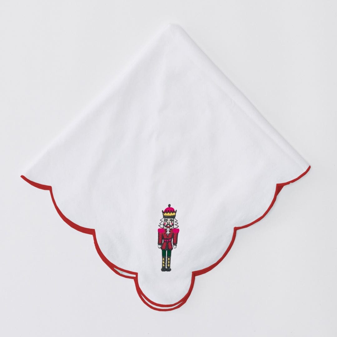 White Nutcracker dinner napkin folded into a point, with a red scalloped edge and embroidered nutcracker, on light gray