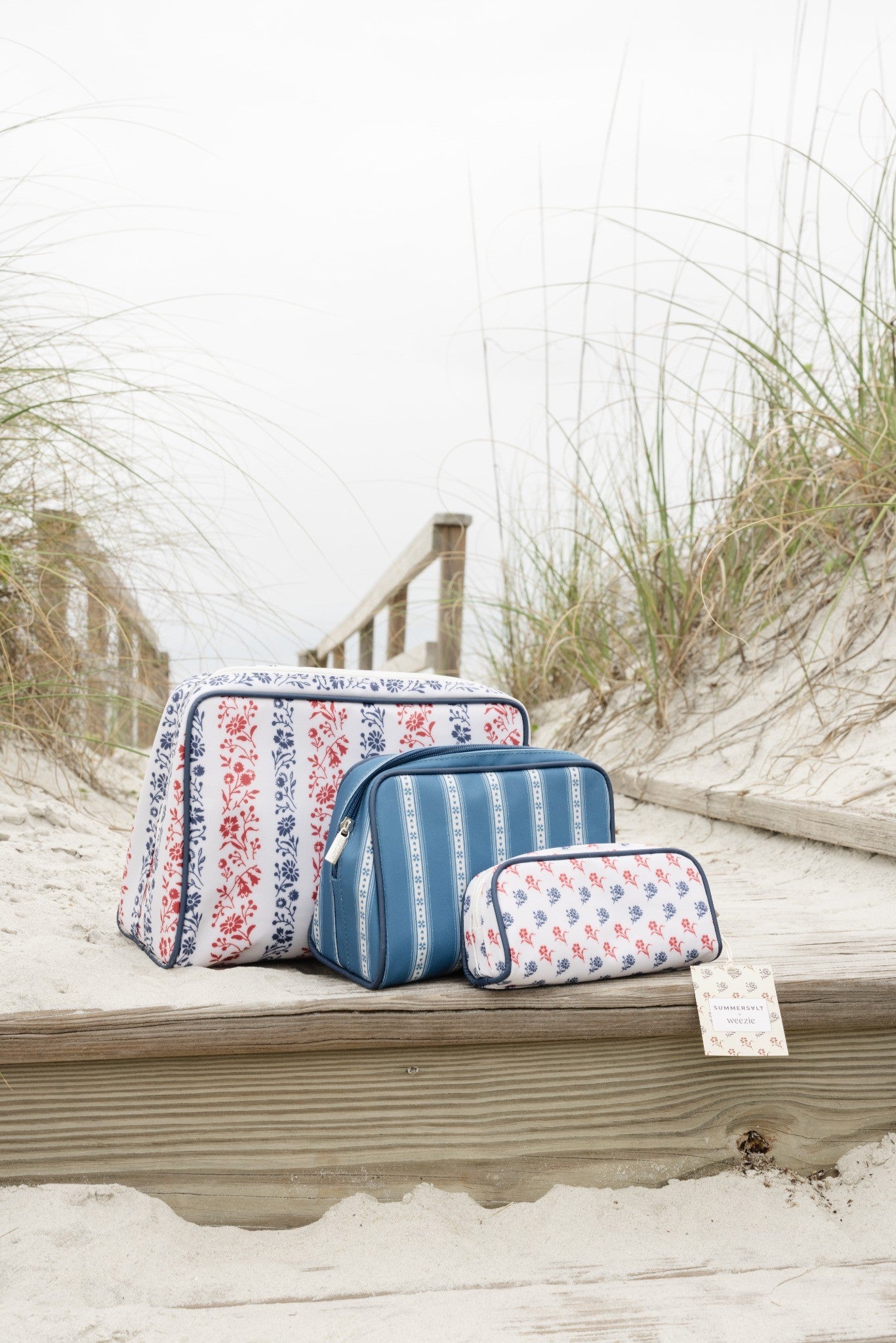 Set of three floral and striped makeup bags on a wooden ledge with sand and grass in the background